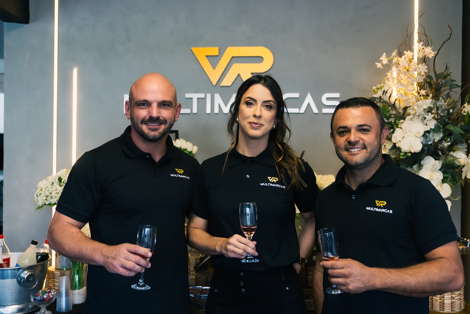 Três sócios da loja com taças na mão em frente ao logo VR Multimarcas